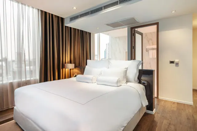A made-up bed with white linen and two bolster pillows sits in a bedroom beside sheer and brown curtains, with a tall building visible through the window and an open doorway to a marble bathroom.