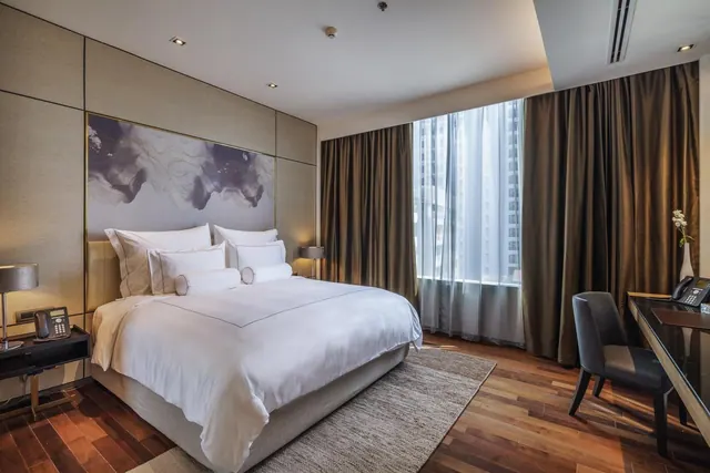 A king bed with white linen sits in a modern hotel bedroom beside a desk, with a large window showing high-rise buildings outside.