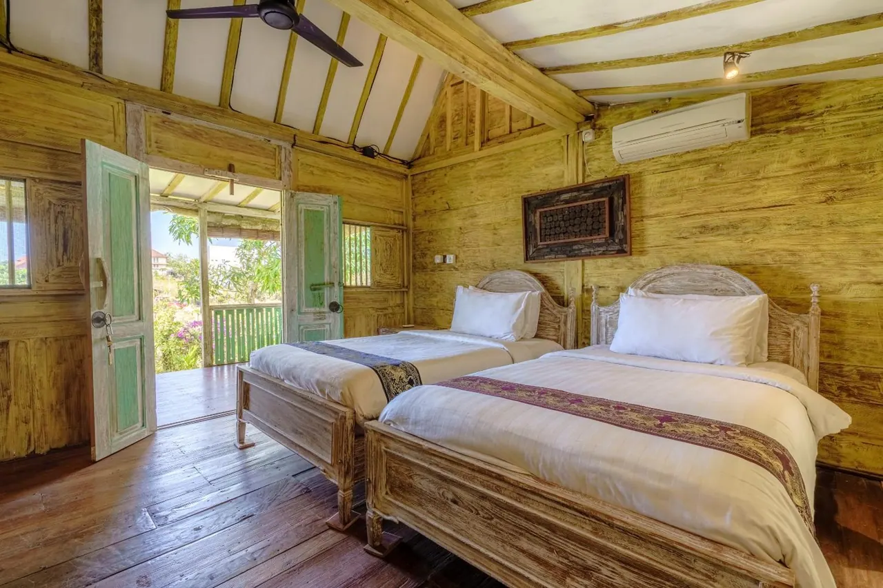 A bedroom with two single beds, a ceiling fan and a wall-mounted air conditioner, with an open door onto a veranda overlooking trees.