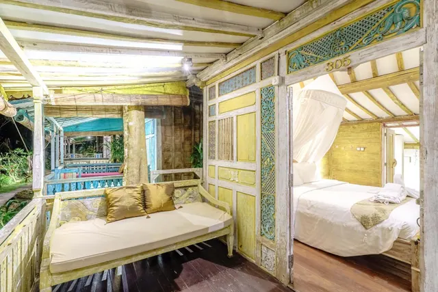 A carved wooden daybed with gold cushions sits on a veranda beside a doorway into a bedroom with a double bed and mosquito net.