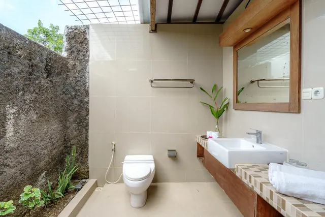 Open-air bathroom with a toilet, a handheld bidet hose, a framed mirror above a single basin on a tiled wooden vanity, and a stone wall on the left.