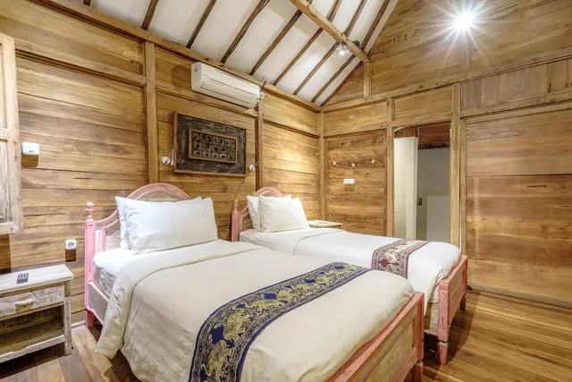 A bedroom with two single beds with pink wooden frames, white bedding and patterned runners, in a room with timber walls, an exposed timber ceiling and a wall-mounted air conditioner.