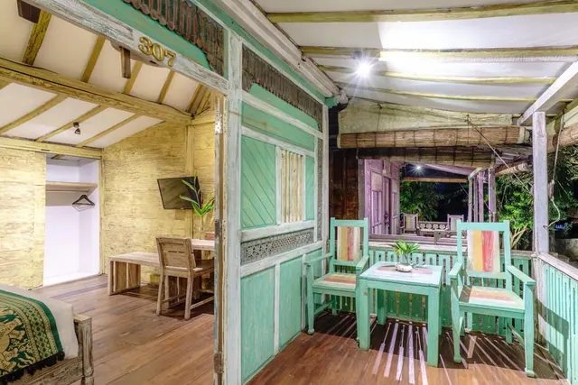 A covered veranda with teal painted chairs and a small table sits beside a room with a bed, wooden desk, wall-mounted television and wardrobe alcove, with trees visible outside at night.