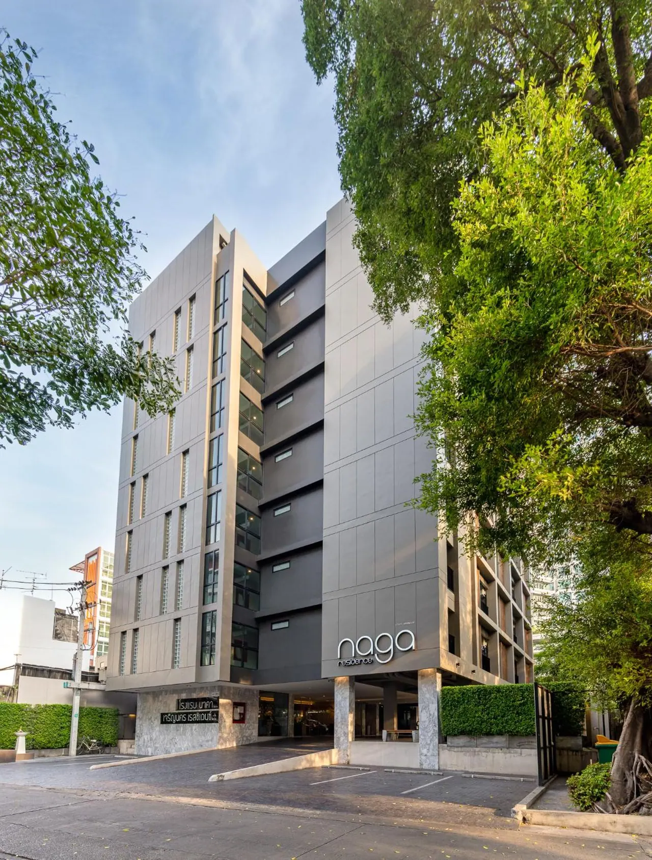 Exterior of a multi-storey grey building with the sign naga residence, a forecourt with parking spaces, and large trees on the right.