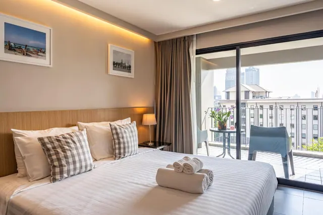 A bedroom with a made bed with striped linen and folded towels, and a glass sliding door opening onto a balcony with chairs and a view of city buildings.