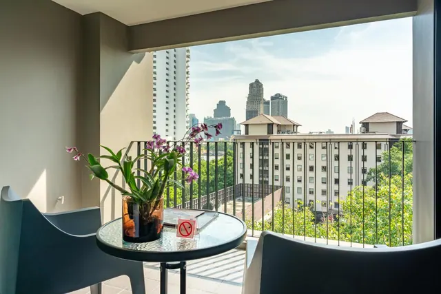 A round glass table with an orchid in a vase and two chairs sit on a balcony overlooking high-rise buildings, trees and towers under a pale sky.