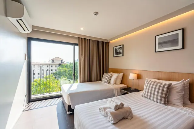 A bedroom with two beds, folded towels on the foreground bed, and a sliding door opening onto a balcony with trees and a building outside.