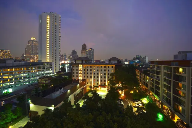 Elevated evening view over a Bangkok cityscape with illuminated high-rise towers, low-rise buildings, trees and parked cars visible below.