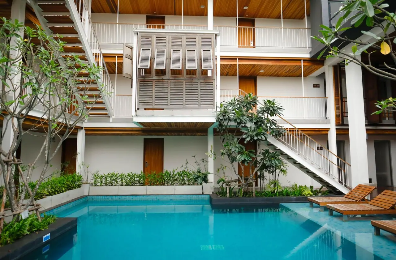 A courtyard swimming pool with wooden sun loungers beside a two-storey white building with timber ceilings, shuttered panels and external staircases.