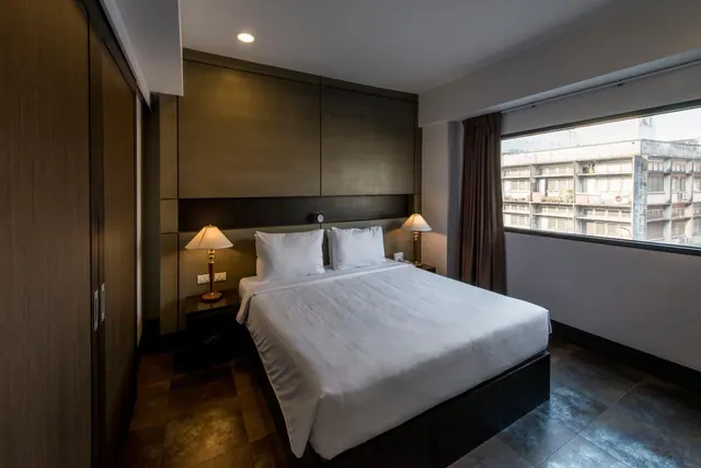 A bedroom with a made king bed, two bedside lamps, a wooden wardrobe and a window looking onto a neighbouring building.