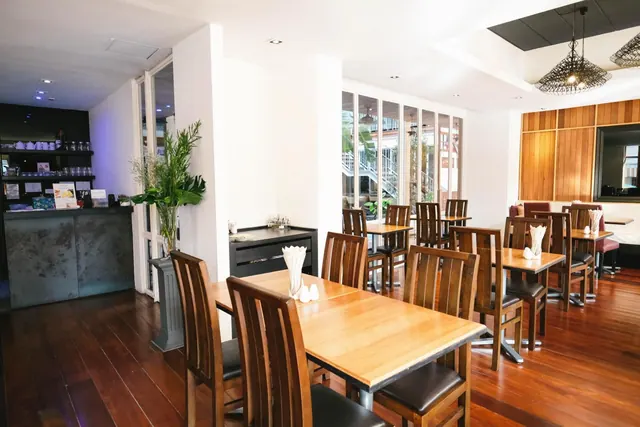 A bright dining room with wooden tables and chairs, a bar counter on the left, and large windows looking onto a staircase and plants outside.