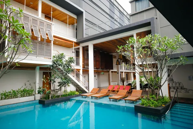 A courtyard swimming pool with wooden sun loungers, frangipani trees, and white colonnaded buildings with balconies around it.