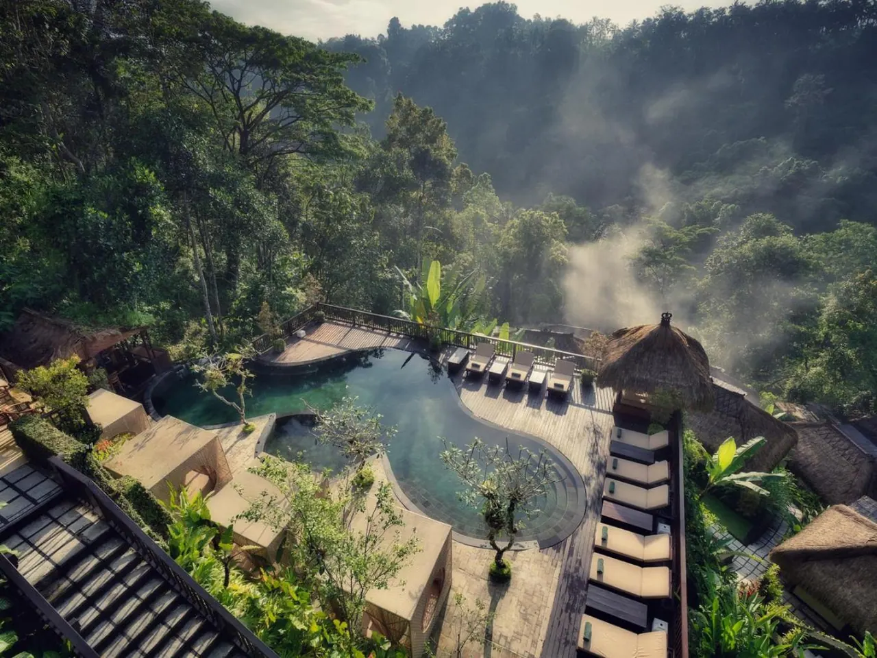Aerial view of a kidney-shaped pool on a timber deck with a row of sun loungers, overlooking a misty forested valley.