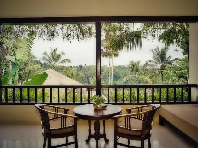 A covered balcony with a round wooden table, two wooden chairs and a daybed, overlooking a green valley of palm trees and a thatched roof.