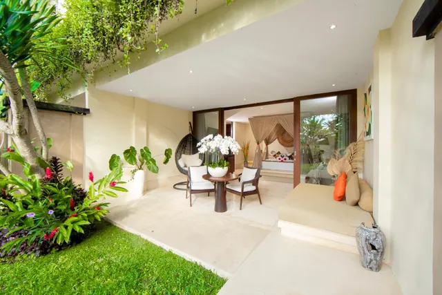 A private outdoor terrace with a round table, two armchairs, a daybed and a view through sliding glass doors into a bedroom with a canopy bed.