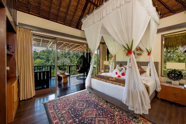 A bedroom with a canopy bed under white netting, a patterned rug, and glass doors opening onto a balcony with a hanging chair and trees beyond.