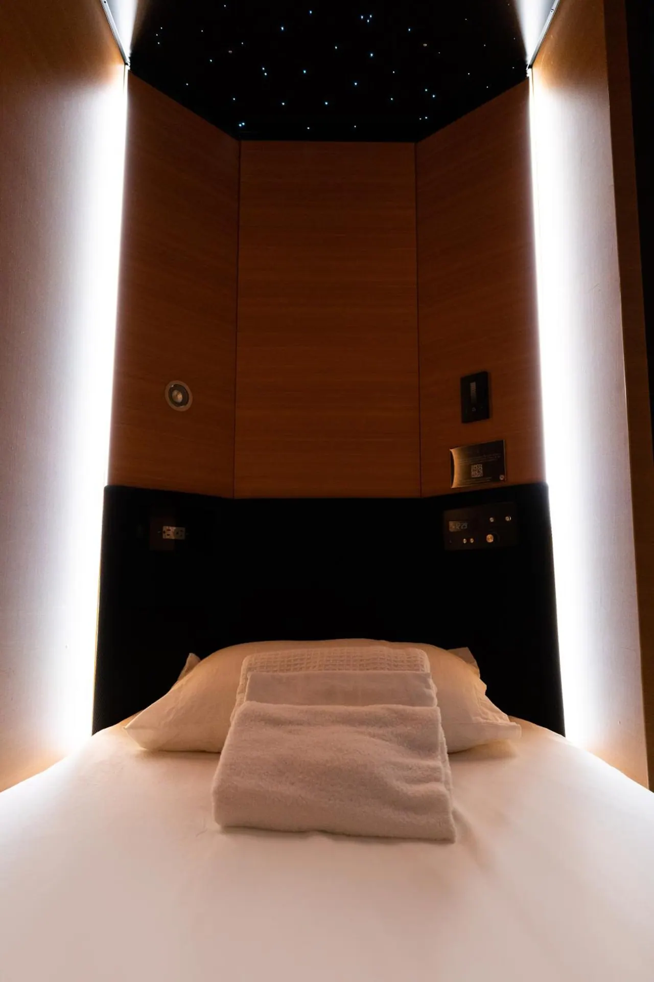 A single bed with white linen and folded towels sits inside a wood-panelled pod with a starry ceiling and edge lighting.