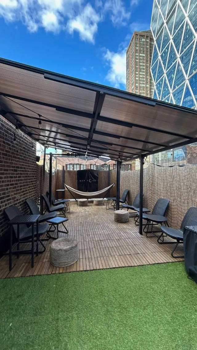 A covered outdoor terrace with wooden decking, artificial grass, wicker lounge chairs, a hammock and stools, with a glass high-rise and a brick wall beside it.