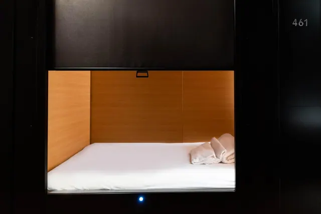 A small enclosed sleeping pod with a made white bed, two folded towels on the mattress, and a small blue light below the opening.