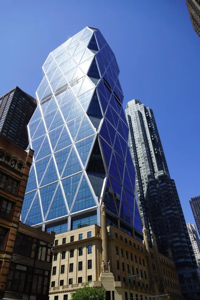 Upward view of the glass-clad Hearst Tower rising above a stone Art Deco base, with neighbouring skyscrapers and a clear blue sky.