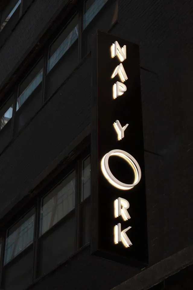 A vertical illuminated sign reading NEW YORK is mounted on a dark building facade at night, with rows of windows beside it.