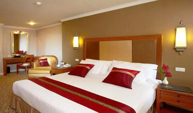 A king bed with white linen, a red runner and red cushions fills a hotel bedroom, with an armchair, a dressing table with mirror and wooden nightstands beside it.