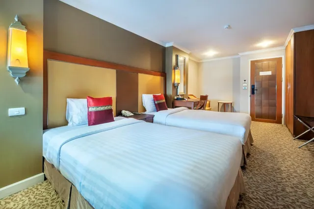A hotel room with two twin beds made with white bedding and red cushions, a wooden desk and chair, and a wooden wardrobe beside the door.