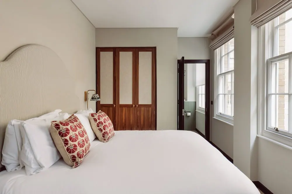 A made double bed with white linen and red patterned cushions stands beside a wooden wardrobe and tall sash windows in a bedroom.