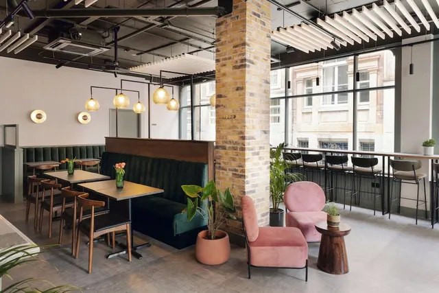 A lounge area with green velvet booths, pink armchairs, wooden dining chairs and a high bar counter by large windows, with an exposed brick pillar in the centre.