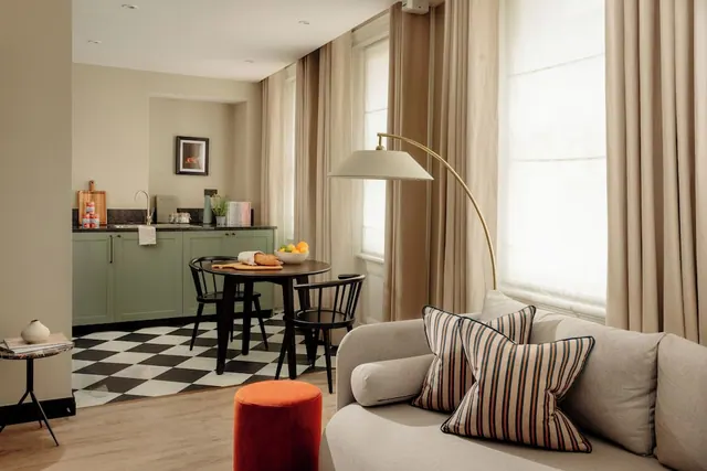 A living area with a grey sofa with striped cushions, a brass arc floor lamp, a round dining table and a green kitchenette on a checkerboard floor.