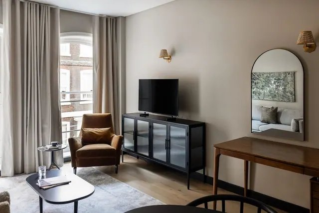 A living area with a brown leather armchair, a glass-top coffee table, a flat-screen television on a black sideboard, and an arched mirror beside a wooden desk, with a brick building visible through the window.