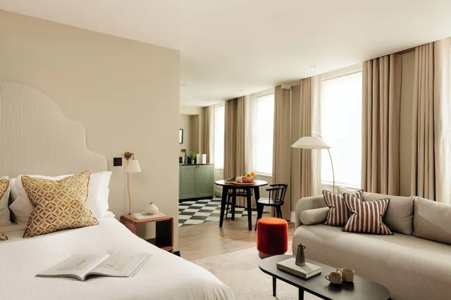 A hotel room with a large white bed in the foreground, a grey sofa with striped cushions, a small round table with chairs, and a green cabinet unit in the background.