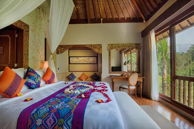 A bedroom with a canopy bed decorated with petals and a woven runner, a wooden desk and chair, and large windows looking onto palm trees and jungle.