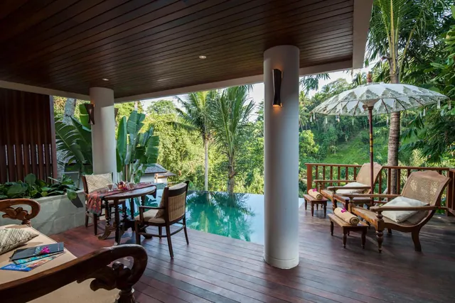 A covered wooden terrace with lounge chairs, a dining table and an infinity pool, with jungle and hillside beyond.