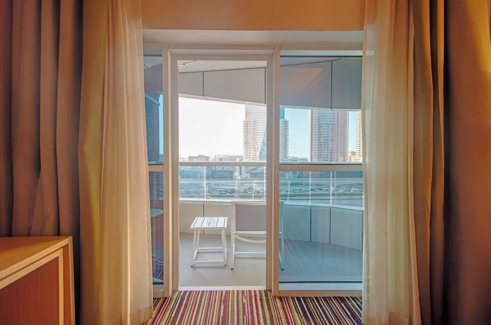 A view through glass doors from a hotel room onto a balcony with a white table and chair, with city towers and a road beyond.