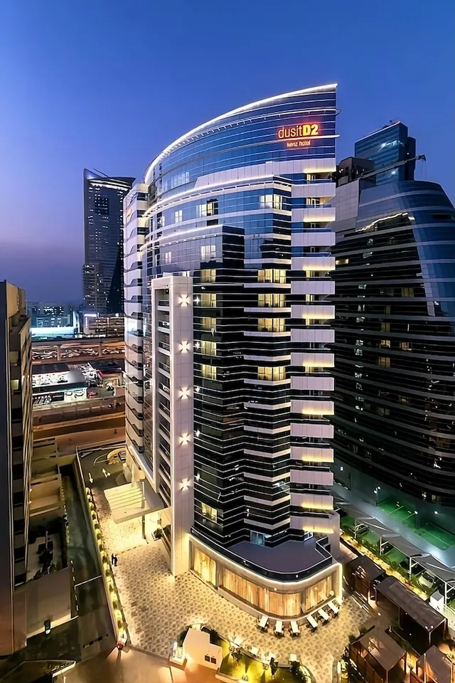 A dusk exterior view of a curved high-rise hotel with illuminated windows and a lit entrance courtyard, with other towers beside it.