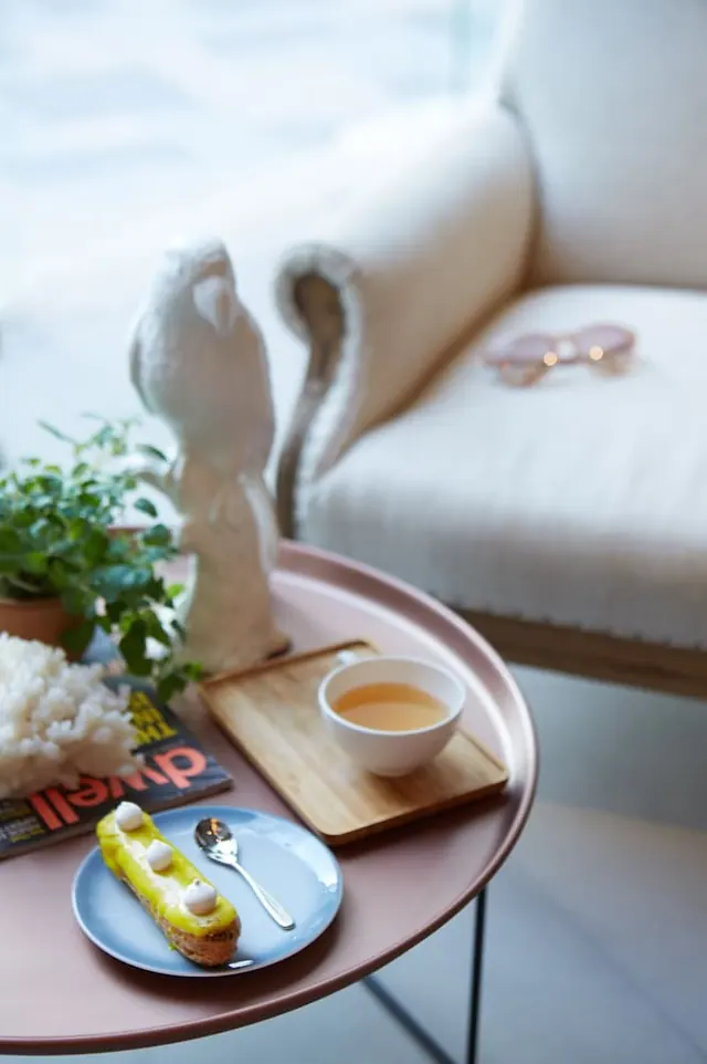A styled side table holds a tea bowl on a wooden tray, an eclair on a blue plate, a magazine, a potted plant and a white bird figurine beside a cream armchair.