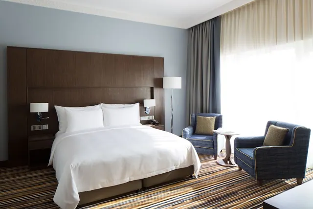 A bedroom with a king bed under white linens, a dark wooden headboard, two blue armchairs and a floor lamp beside a large window with sheer curtains.