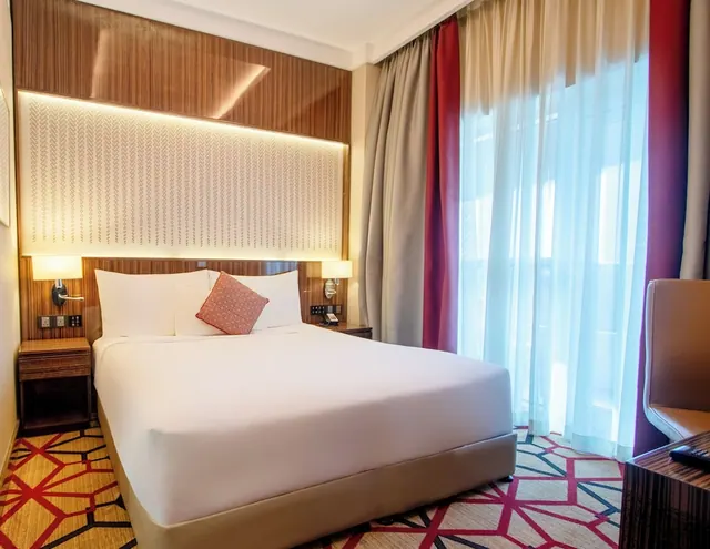 A bedroom with a made double bed, a red patterned cushion, a wood-panelled headboard wall with a backlit panel, and sheer curtains over a large window.
