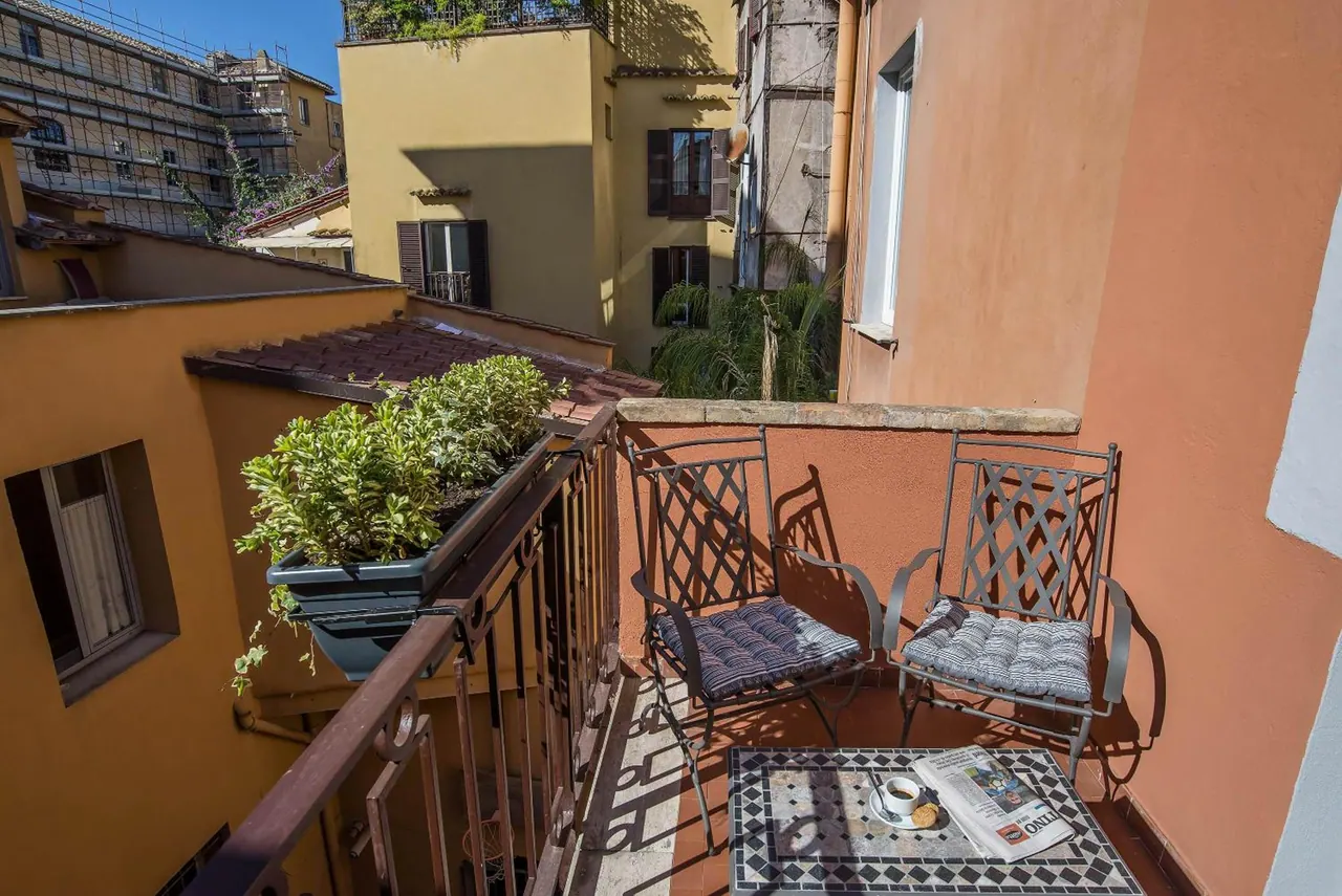 A private balcony with two wrought-iron chairs, a mosaic-topped table holding a coffee cup and newspaper, and a potted plant on the railing, looking out over neighbouring buildings.