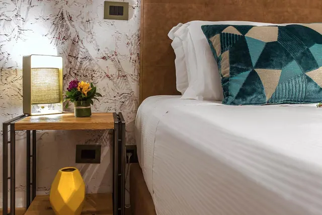 A bedside table with a lamp, a small flower arrangement and a yellow vase stands beside a made bed with white linen and a teal patterned cushion.