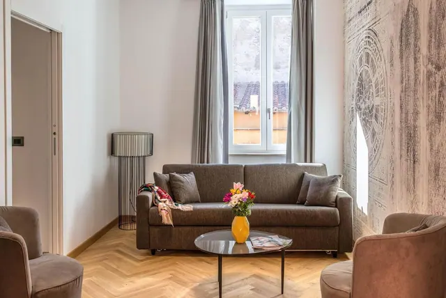 A living area with a brown sofa, a round glass coffee table holding a yellow vase of flowers, two armchairs, and a window looking onto a neighbouring building with tiled roofs.
