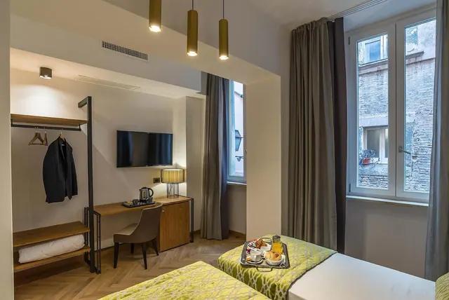 A hotel bedroom with two single beds, one holding a breakfast tray, a wall-mounted television, a desk with a kettle, and a clothing rail, with a window looking onto a brick wall.