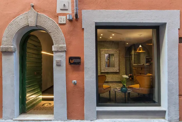 Street-level facade of a hotel with an arched stone doorway, a partly open green door, and a large window showing a lounge with armchairs, a small table and a pendant lamp.
