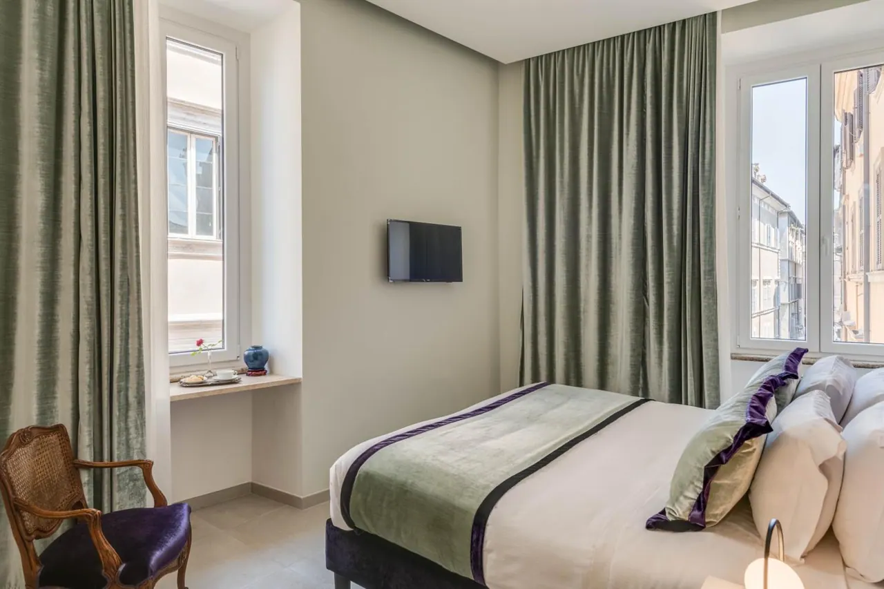 A bedroom with a double bed dressed in white linen, green velvet curtains, a wall-mounted television and a cane armchair, with windows looking onto neighbouring buildings across a street.