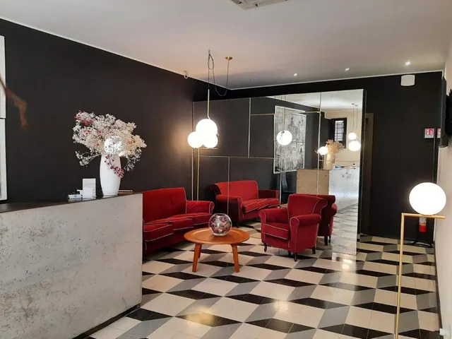 A lobby with red armchairs and a red sofa around a round wooden table, a checkered floor, a concrete reception desk, and a white vase of dried flowers.