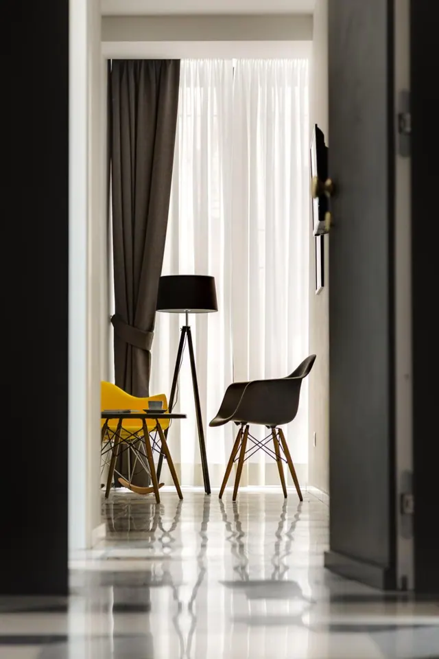A seating area with a tripod floor lamp, a yellow chair, a dark armchair and a small table, seen through a doorway with sheer curtains behind.