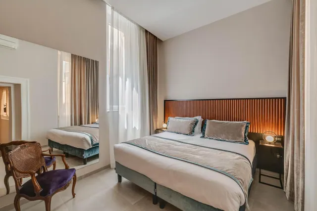 A bedroom with a double bed under a slatted wooden headboard, a wall mirror reflecting the bed, and two cane-back chairs with purple velvet seats by a sheer-curtained window.