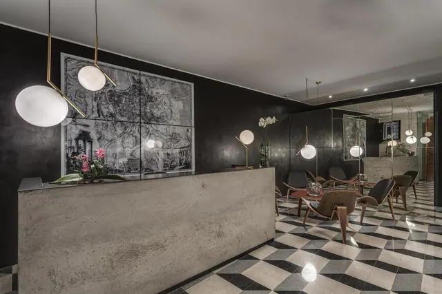 A lobby with a concrete reception desk, black walls, a framed map of Rome, pendant globe lights, and lounge chairs on a checkered tiled floor.
