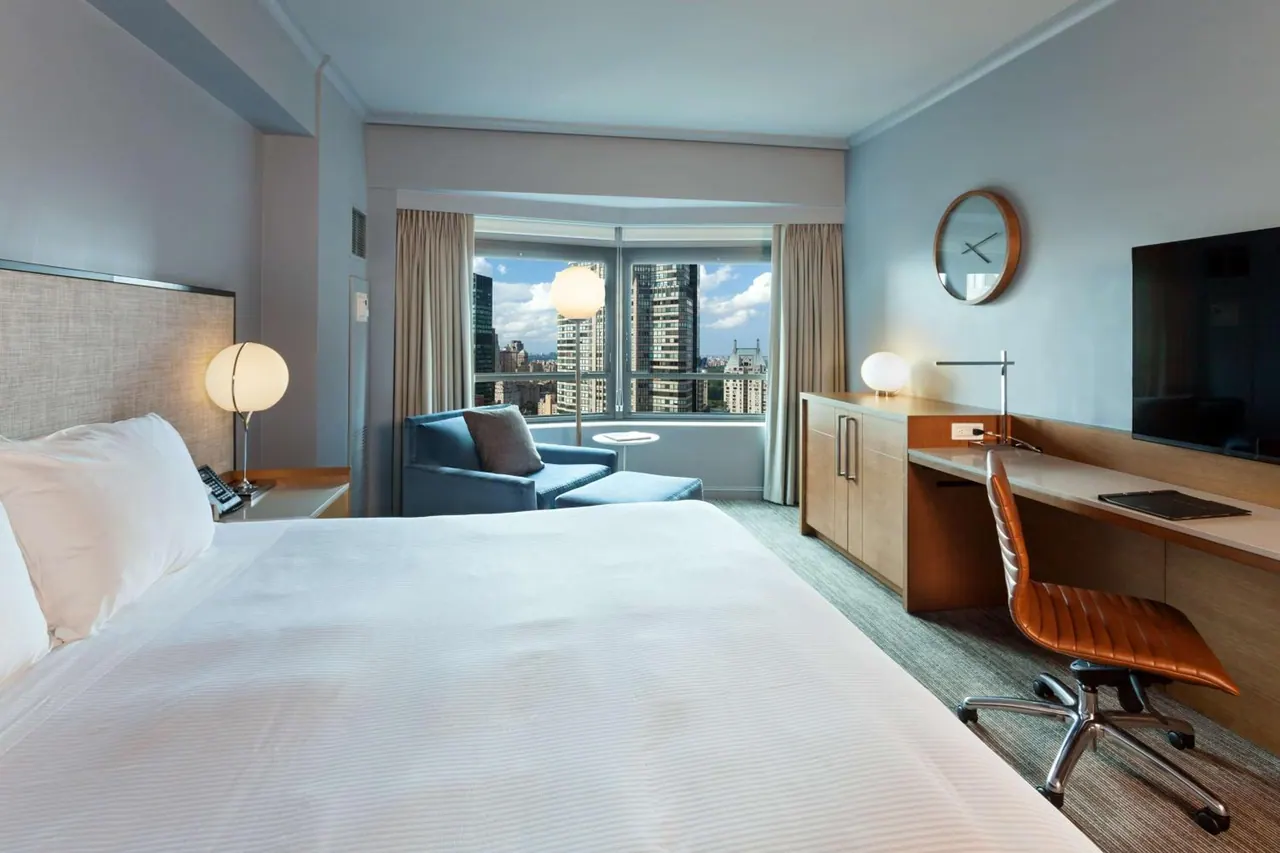 A hotel bedroom with a large white bed, a blue armchair by a window overlooking city towers, a wooden desk with a chair, and a wall-mounted television.